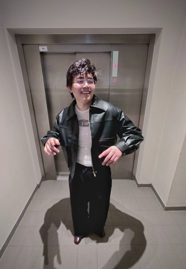 Person med kort krøllet hår og briller, iført svart skinnjakke og mørke bukser, står og smiler foran lukkede heisdører.
