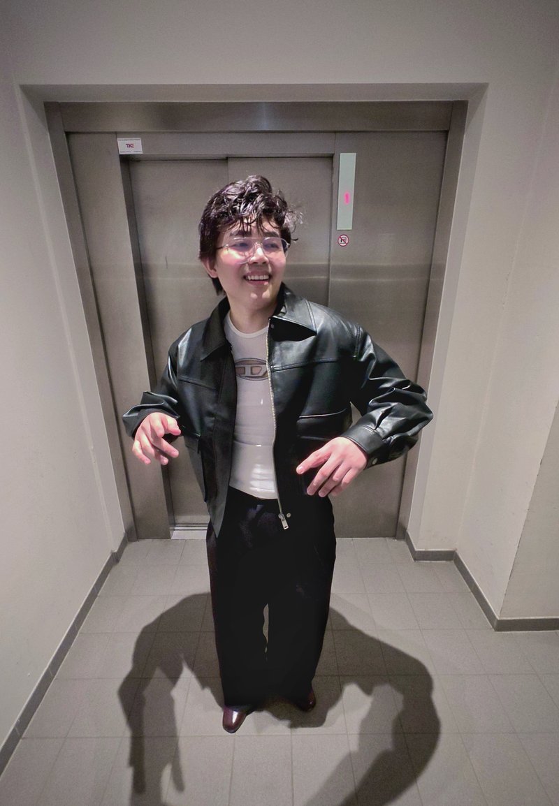 Personne aux cheveux courts et bouclés portant des lunettes, vêtu d'une veste en cuir noire et d'un pantalon foncé, debout et souriant devant des portes d'ascenseur fermées.