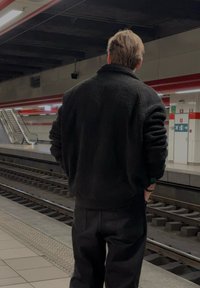 Purūs juodi pūkų džemperis, juodos kelnės, stovintis metro stotelėje, fone matomi eskalatorius ir bėgiai.