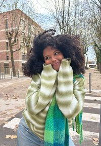 Πράσινο και κρεμ ριγέ πουλόβερ με χαλαρούς ώμους, συμπληρωμένο από ένα κασκόλ σε καρό μπλε και πράσινο. Υπαίθρια σκηνίτσα με δέντρα και τούβλινο κτίριο.