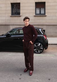 Bordowy sweter, dopasowane spodnie i burgundowe mokasyny. W tle czarny samochód, teksturowana ściana budynku oraz dwa okna.