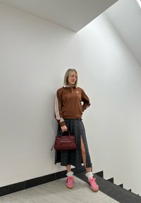 Hanorac Adidas marron cu dungi albe, fustă scoasă cu carouri negre și o fanta, adidași roz și o geantă marsala, stând în fața unui perete alb.