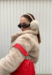 Ψεύτικο γούνινο παλτό σε κρέμα, μαύρα γυαλιά ηλίου, λευκά ακουστικά και μια κόκκινη τσάντα με υφή και έντονο λογότυπο σε αντίθετο λουράκι.