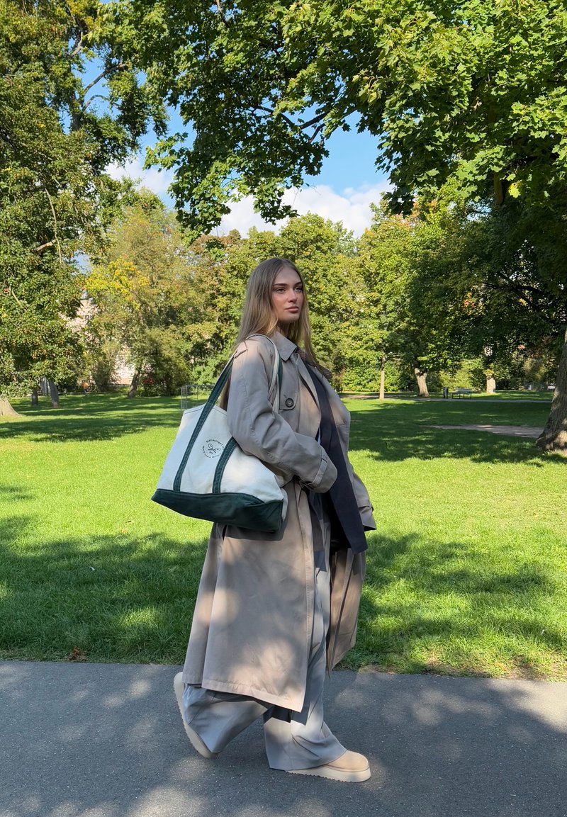 Trench coat bege, calças cinzentas largas e sapatos bege. Top preto e mala de ombro com detalhes verdes e logótipo. Caminha num caminho de um parque.