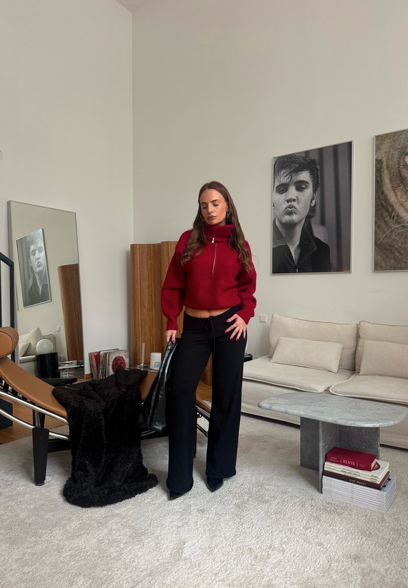 Piros rövid pulóvert és fekete nadrágot viselő nő egy modern nappaliban áll, melyben bézs kanapé, fekete szék és keretezett fekete-fehér portrék találhatók.