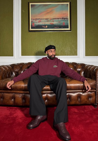 Sudadera marrón con logo verde, pantalones anchos negros y mocasines marrones oscuros. Sentado en un sofá de cuero marrón tapizado contra una pared verde.