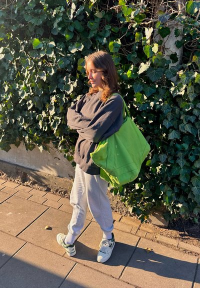 Joven mujer vestida de manera casual con los brazos cruzados, de pie en la acera junto a una pared cubierta de hiedra, llevando una gran bolsa de tela verde.
