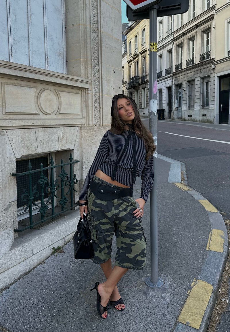 Jeune femme en haut rayé et short camouflage pose sur le trottoir de la ville près d'un panneau de signalisation, tenant un sac à main noir et portant des talons noirs.