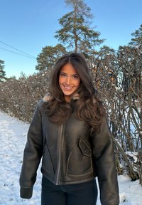 Jeune femme souriante, portant une veste en cuir noire, debout sur un chemin enneigé entouré de buissons sans feuilles et de pins sous un ciel bleu clair.