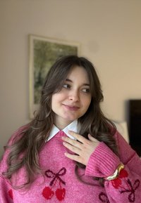 Vaaleanpunainen neulepaita kirsikkakuvioilla, alla valkoinen kauluksellinen paita, pitkät hihat resorilla varustettuina, käytettynä kultainen rannekoru.