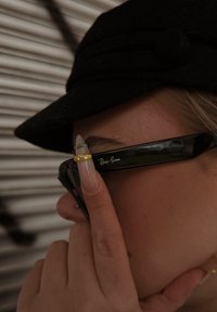 Fekete Ray-Ban napszemüveg fényes felülettel; a logó látható a kereten. Egy kéz, hosszú, díszes körmökkel, a szemüveg közelében helyezkedik el.