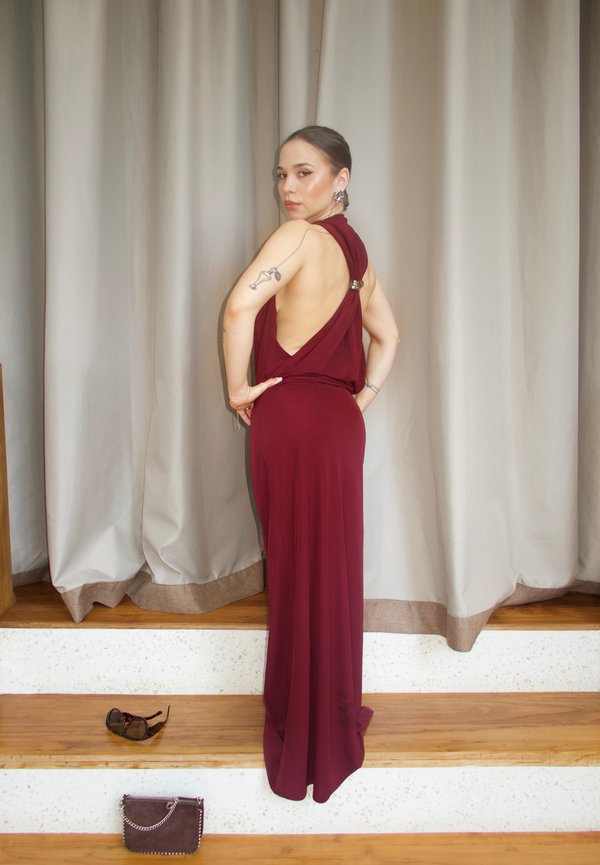 Nainen hihattomassa burgundin värisessä selkämyksettömässä mekossa seisoo puuportailla aurinkolasit ja laukku lähellä, katsoo olkapään yli.