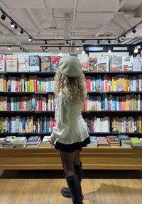 Una donna è in piedi vicino a una libreria nera piena di libri colorati. Indossa una camicetta bianca, una gonna nera e stivali neri al ginocchio.