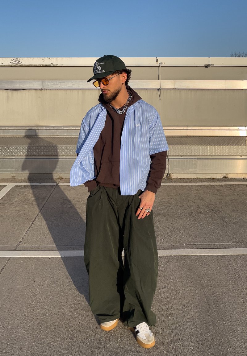 Jeune homme portant une casquette noire, des lunettes de soleil orange, une chemise à rayures bleues superposée sur un sweat à capuche marron, un pantalon ample vert foncé et des baskets blanches sur le trottoir.