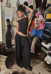 Young woman in black outfit adjusting her hair, standing in a cluttered room with clothes, boots, and bags on shelves and hooks.