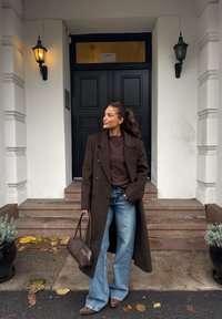 Donna con cappotto marrone e maglione, jeans blu e stivali marroni, in piedi con una borsa abbinata sui gradini davanti a una porta nera con pareti bianche.