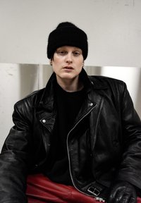 Veste de motard en cuir noir, bonnet en maille noire, chemise noire unie et pantalon en cuir rouge. Les accents comprennent des éléments en métal argenté et une variation de textures.