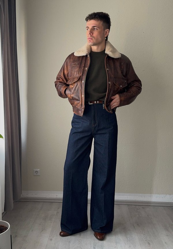 Jauns vīrietis, tērpies brūnā ādas jaku ar kažokādas apkakli, tumši zaļu džemperi, plašām tumšām džinsa biksēm un brūnām kurpēm, stāv iekštelpās pie loga.