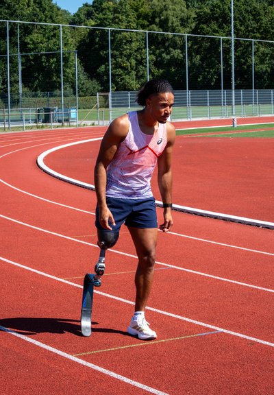 Atleta con una prótesis de hoja para correr en una pista roja, vestido con una camiseta sin mangas y pantalones cortos, preparándose para correr bajo un cielo soleado.