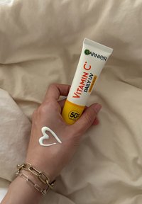 Tubetto di crema giornaliera UV alla vitamina C di Garnier, bianco con accenti gialli, tenuto in mano. Texture cremosa sulla pelle che forma una forma a cuore.