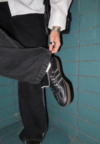 Melns ādas sporta apavu ar baltu šuvumu, gumijas zoli un zelta akcentiem; valkāts kopā ar melnām džinsa biksēm. Fonā redzamas zaļganas flīzētas sienas.