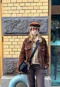 Hnědá semišová bunda s bílým prošíváním, béžový svetr, hnědé pruhované kalhoty a hnědá čepice; velké čtvercové sluneční brýle a kabelka přes rameno.