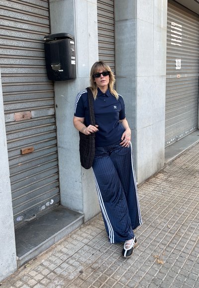 Persona vestida con una polo navy de Adidas y pantalones anchos a rayas se apoya contra un pilar de piedra junto a persianas metálicas cerradas en una acera urbana.