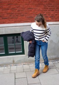 Suéter a rayas blanco y marino, chaqueta acolchada oscura, jeans azules y botas de cordones color canela con cordones amarillos, de pie en una acera adoquinada.