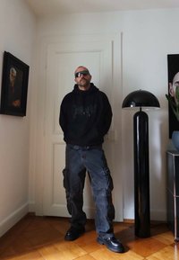 Felpa nera con cappuccio e grafica grigia, pantaloni cargo scuri con multiple tasche e scarpe nere. In piedi al chiuso accanto a una lampada alta nera.