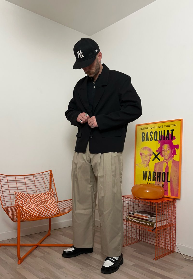 Homme en blazer noir et pantalon large beige boutonnant sa veste, portant des chaussures noires et blanches ainsi qu'une casquette noire des New York Yankees dans une pièce moderne.