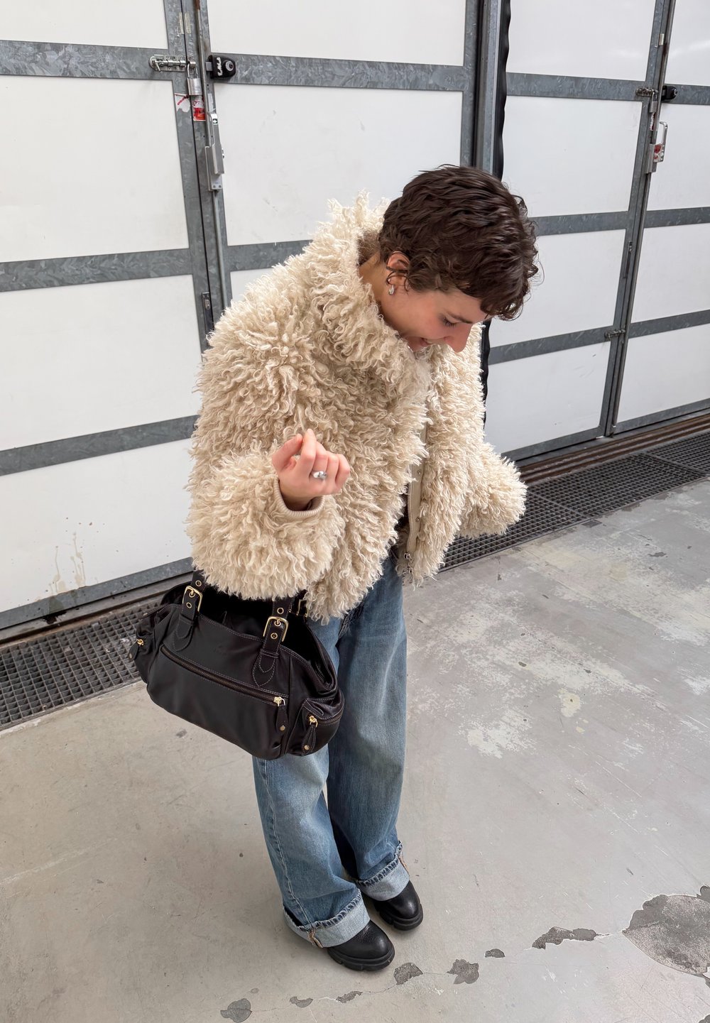 Personne portant une veste beige duveteuse et un jean, tenant un sac à main noir, regardant vers le bas et debout sur un sol en béton près de portes de garage en métal.