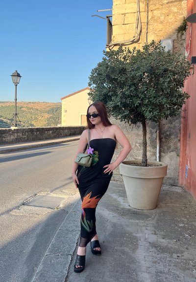 Vestido negro sin mangas con estampado floral, combinado con tacones de plataforma negros. Pequeño bolso verde con cadena y detalle de flor morada.