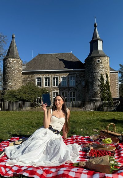 Vestido blanco con tejido texturizado, cinturón negro y perlas, sentada sobre una manta a rayas rojas y blancas, rodeada de frutas y una cesta de picnic.