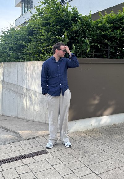 Conjunto informal que incluye una camisa de botones azul oscuro y pantalones beige claros. Zapatillas deportivas grises complementan el look. Entorno urbano con vegetación.