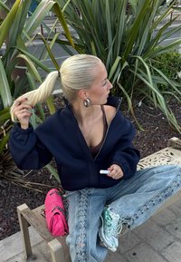 Žena s plavim repom nosi mornarsku jaknu, traperice s čipkastim vezicama i tenisice te sjedi na klupi, držeći bijeli cilindrični predmet u blizini zelenog bilja.