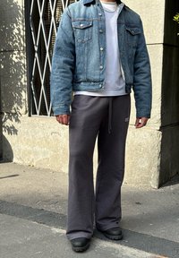 Chaqueta de mezclilla con lavado azul, hombros ajustados y dos bolsillos en el pecho. Camiseta blanca debajo. Pantalones de chándal grises sueltos y zapatos negros.