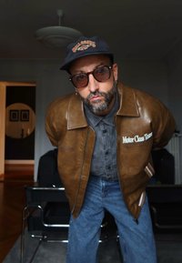 Mann iført en brun skinnjakke, grå skjorte, blå jeans, solbriller og svart caps som lenes fremover innendørs med en mørk bakgrunn.