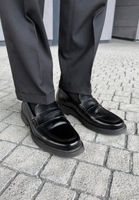 Sorte læderloafers med sorte sokker og mørke bukser, som står på grå stenfliser udendørs.