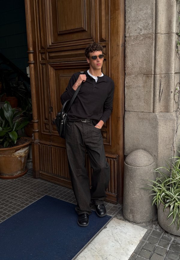 Homme en lunettes de soleil et vêtements sombres appuyé contre une grande porte en bois, tenant un sac noir, avec des plantes et des murs en pierre à proximité.