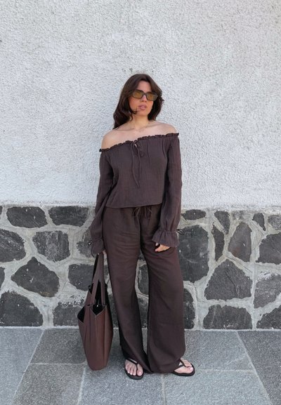 Top marrón sin mangas y pantalones de pierna ancha a juego, hechos de un tejido ligero, con bordes con volantes y un cordón. Bolsa tote marrón y chanclas.