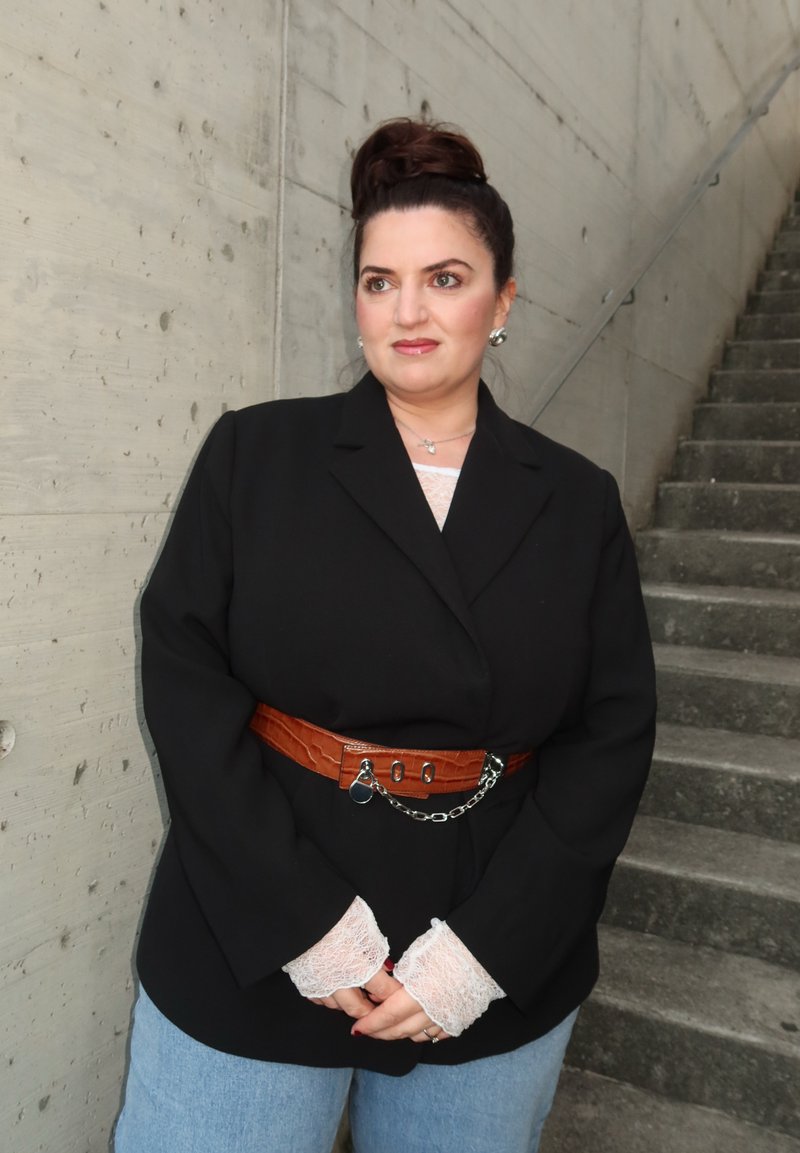 Femme aux cheveux foncés attachés en chignon, portant un blazer noir, une ceinture marron avec chaîne, des poignets en dentelle et un jean bleu clair, se tenant près d'escaliers en béton.