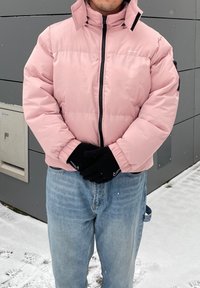 Casaco acolchoado rosa com gola alta, fecho preto e bolsos laterais; usado sobre jeans de denim azul e luvas pretas; fundo nevado.