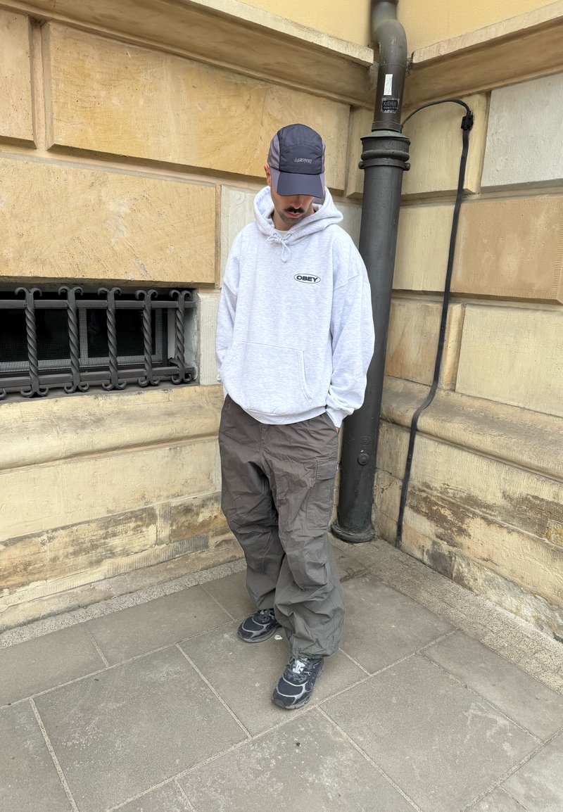 Homme en sweat à capuche gris, pantalons cargo foncés, baskets et casquette debout sur le trottoir de la ville près d'un bâtiment en pierre beige et d'une fenêtre avec grille métallique noire.