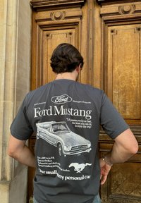 Γκρι βαμβακερό μπλουζάκι με γραφικό vintage Ford Mustang, κείμενα λεπτομέρειες σχετικά με το αυτοκίνητο και μια σιλουέτα ενός καλπάζοντος αλόγου.