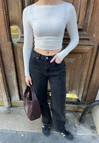 Witte lange mouwen cropped top, hoge taille zwarte spijkerbroek, donkere sneakers en een bruine handtas, staand tegen een houten deur.