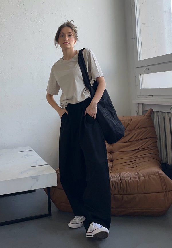 Beige t-shirt, loose black trousers, and a black tote bag. White trainers. Simple design, relaxed fit, smooth fabric, and minimal accents.