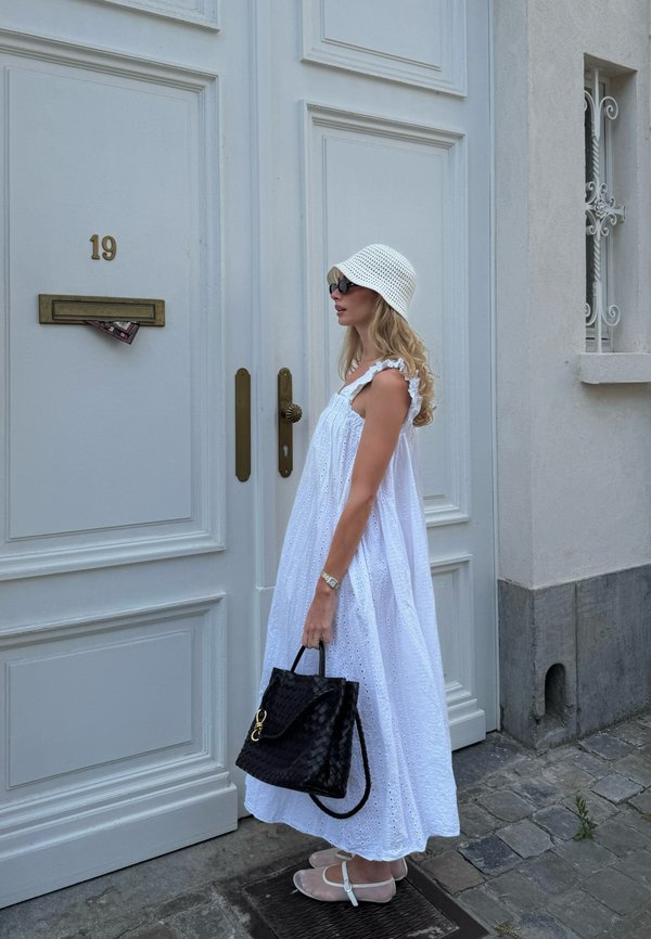 Woman in white eyelet dress and hat holding black handbag stands beside white door with number 19 and mail in letterbox.