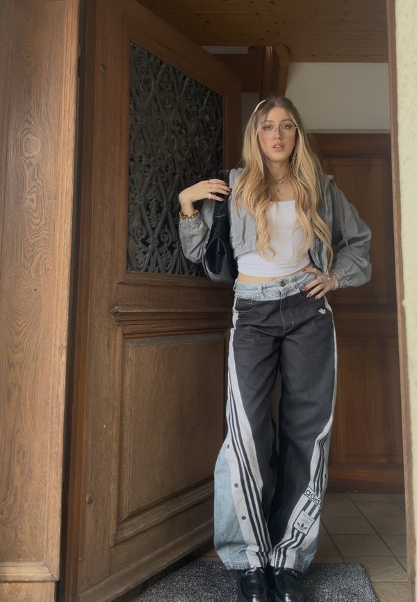 Tânără cu păr lung și blond purtând ochelari, jachetă scurtă, top alb și pantaloni largi Adidas, stând în cadrul unei uși din lemn.