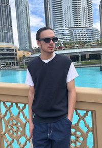 Joven con gafas de sol, camisa blanca debajo de un chaleco negro, de pie junto a una barandilla ornamentada cerca del agua azul, con rascacielos modernos de fondo.