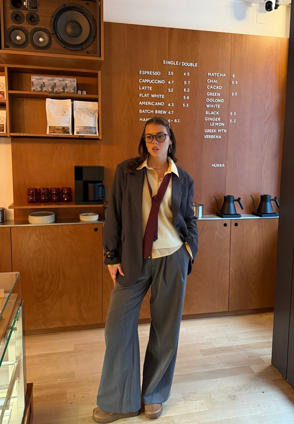Jeune femme portant un blazer oversize, un pantalon ample et des lunettes, debout devant le menu en bois d'un café affichant les options de café et de thé.